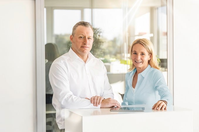 Ein Mann und eine Frau stehen an einem modernen weißen Empfangstresen in einem hellen Büro, beide lächeln freundlich
