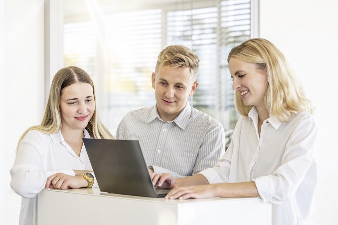 Zwei Frauen und ein Mann stehen zusammen an einem Laptop und arbeiten konzentriert in einem hellen Büro