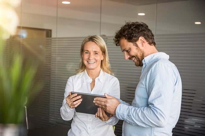 Eine Frau und ein Mann schauen gemeinsam auf ein Tablet und lächeln, in einem modernen Büro mit Glaswänden